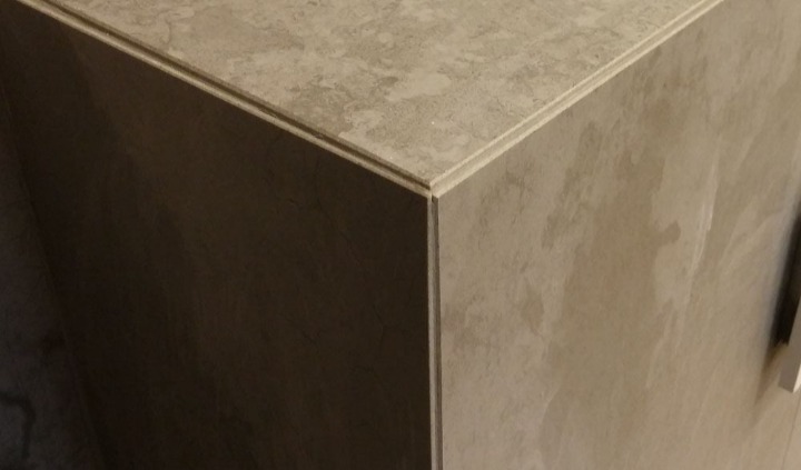 Fragment narożnika obudowy z płytek imitujących kamień, z widocznymi delikatnymi wzorami i jasnymi fugami, oświetlony bocznym światłem.