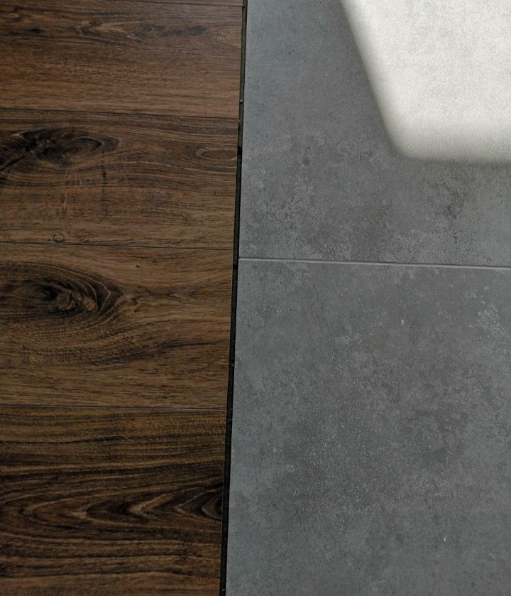Połączenie paneli podłogowych imitujących ciemne drewno i szarej, matowej płytki ceramicznej podłogowej, widoczne fugi i delikatny cień.