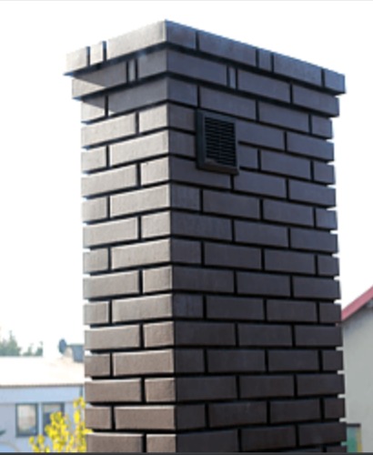 Brązowy komin z cegły klinkierowej z zamontowaną kratką wentylacyjną, widoczny fragment dachu i zieleni w tle.