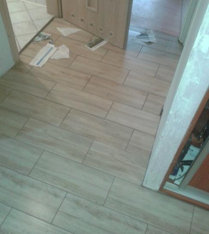 Ułożona podłoga z płytek imitujących drewno, widoczne resztki po montażu w postaci papieru i okruchów, częściowo otwarta drewniana ościeżnica drzwiowa.