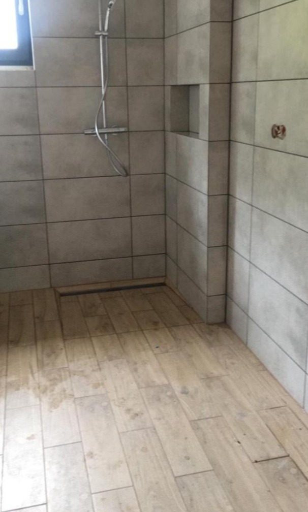 Nowoczesna kabina prysznicowa z szarymi płytkami ściennymi i podłogą imitującą drewno, widoczny odpływ liniowy i wnęka w ścianie. Ślady po budowie na podłodze.