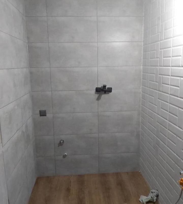 Wnętrze kabiny prysznicowej z szarymi, prostokątnymi płytkami na ścianach, drewnopodobną podłogą i czarną baterią prysznicową, widoczne przyłącza wodne i fragment instalacji.