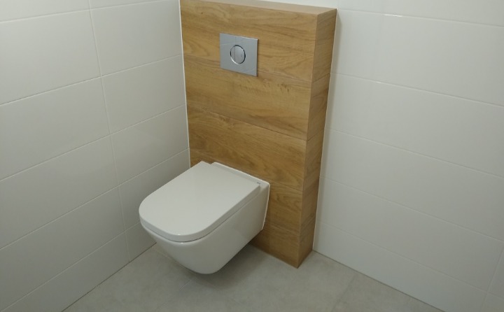 Podwieszana toaleta z drewnianą zabudową i chromowanym przyciskiem spłukującym na tle białych, prostokątnych płytek ściennych i szarych płytek podłogowych.