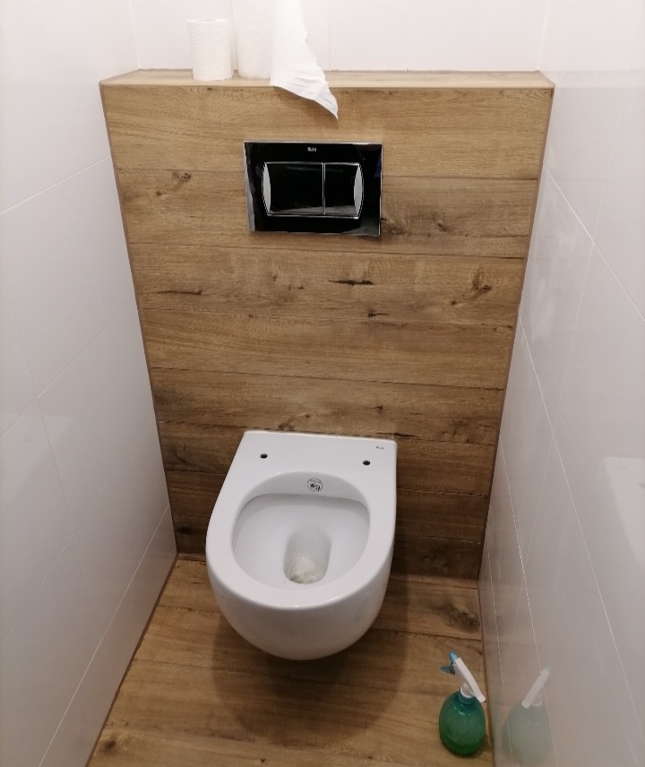 Wisząca miska WC zamontowana na drewnopodobnej ściance z chromowanym przyciskiem spłukującym, białe płytki na ścianach, podłoga w kolorze drewna, butelka ze spryskiwaczem w rogu.