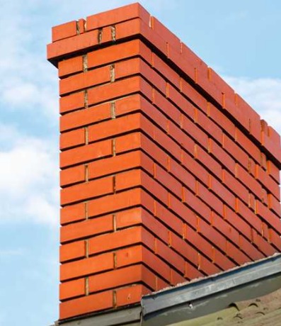 Ceglany komin z widoczną strukturą i spoinami na tle błękitnego nieba, fragment dachu z rynną w dolnej części kadru.