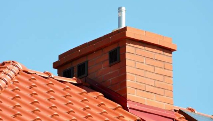 Czerwony dach z ceramicznej dachówki z ceglanym kominem z metalowym wkładem kominowym i kratkami wentylacyjnymi, na tle błękitnego nieba.