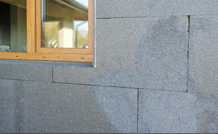 Szara ściana budynku ocieplona styropianem grafitowym, widoczna drewniana rama okienna, fuga między płytami styropianowymi wypełniona pianką.