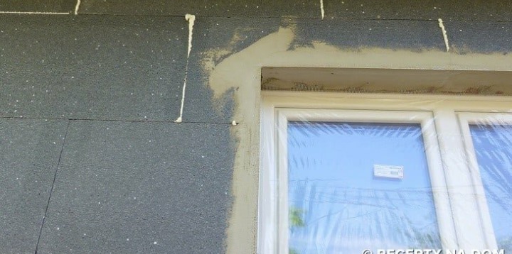 Szara ściana budynku w trakcie ocieplania styropianem, widoczne spoiny wypełnione pianką montażową oraz obramowanie okna z nałożoną warstwą kleju z siatką.