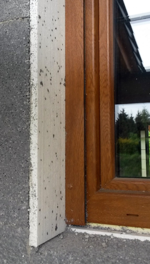 Fragment ocieplonej ściany z zamontowanym oknem o drewnianej ramie, widoczne pozostałości zaprawy i styropianu.