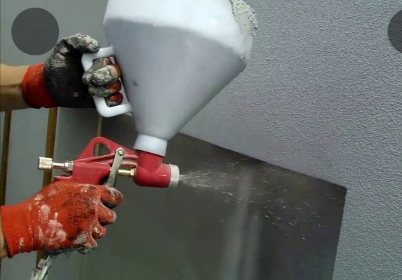 Natryskowa aplikacja tynku strukturalnego na ścianę za pomocą pistoletu z zasobnikiem, widoczne ochronne rękawice robocze.