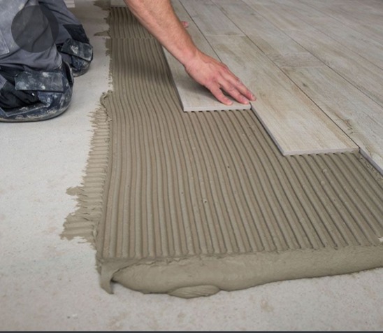 Układanie podłogi z płytek imitujących drewno na świeżo nałożonym kleju z wyraźnymi liniami, widoczne dłonie glazurnika i fragment jego ubrania.