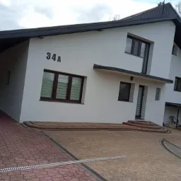 Nowoczesny dom jednorodzinny z jasną elewacją, ciemną stolarką okienną i dachem, widoczny z poziomu brukowanego podjazdu z numerem 34A na ścianie.