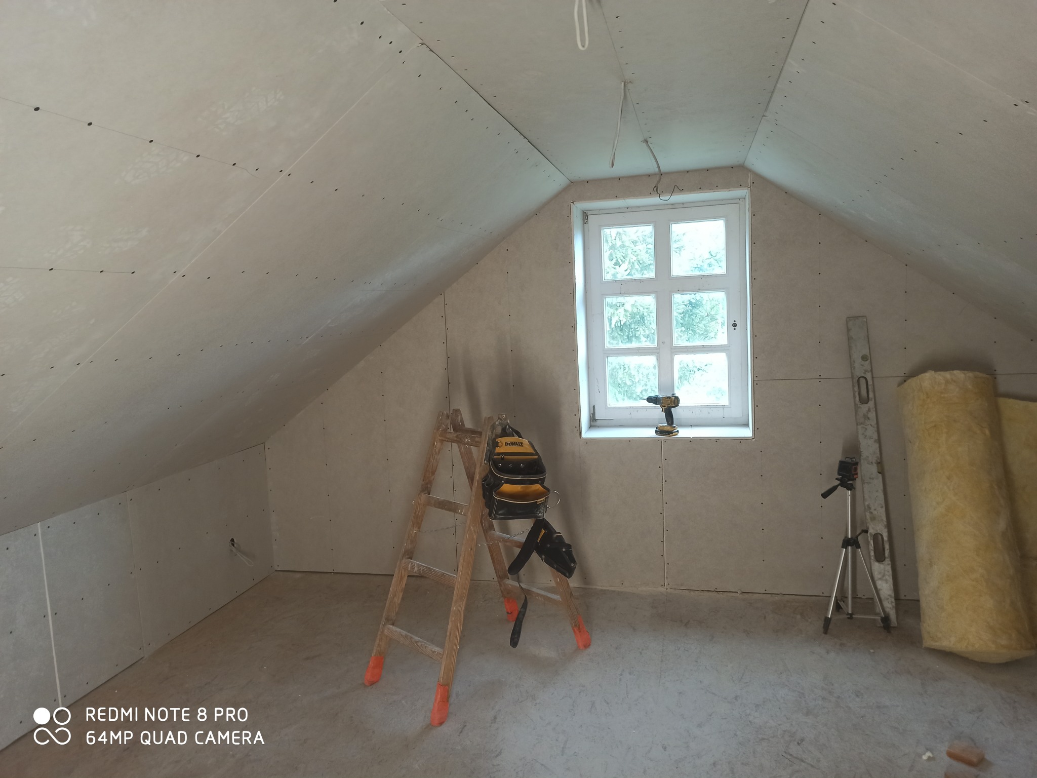 Poddasze w Namysłowie w trakcie wykańczania, ściany pokryte płytami gipsowo-kartonowymi, drabina z narzędziami, poziomica i rolka wełny mineralnej widoczne w pomieszczeniu.
