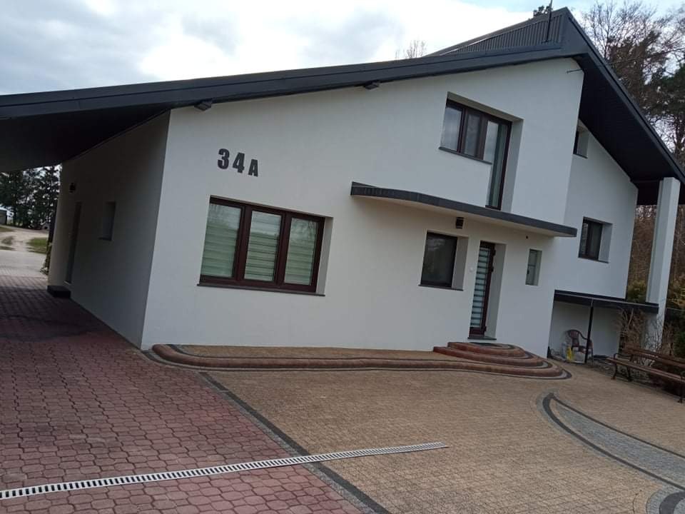 Nowoczesny dom jednorodzinny z jasną elewacją, ciemną stolarką okienną i dachem, widoczny z poziomu brukowanego podjazdu z numerem 34A na ścianie.
