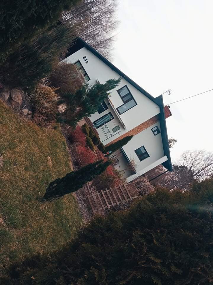 Biały dom z numerem 34A, częściowo otynkowany na brązowo, widziany z perspektywy ogrodu z iglakami i trawnikiem.