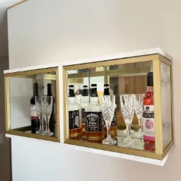 Dwie witryny wiszące z marmurowym blatem i złotymi ramami, eksponujące butelki alkoholu i kryształowe kieliszki, zamontowane na białej ścianie.