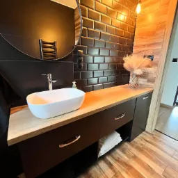 Nowoczesna łazienka z blatem drewnianym, umywalką nablatową i czarnymi płytkami w stylu metro. Uwagę przykuwają lampy wiszące i drewniane wykończenie obok drzwi.