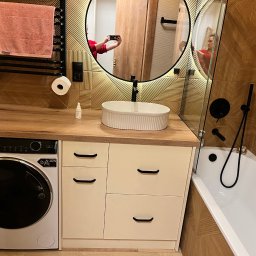 Michał Lodarski - Nowoczesna łazienka z drewnianymi akcentami: umywalka nablatowa, pralka pod blatem, lustro w okrągłej ramie i mozaika na ścianie.