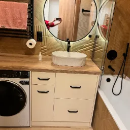 Nowoczesna łazienka z drewnianymi akcentami: umywalka nablatowa, pralka pod blatem, lustro w okrągłej ramie i mozaika na ścianie.