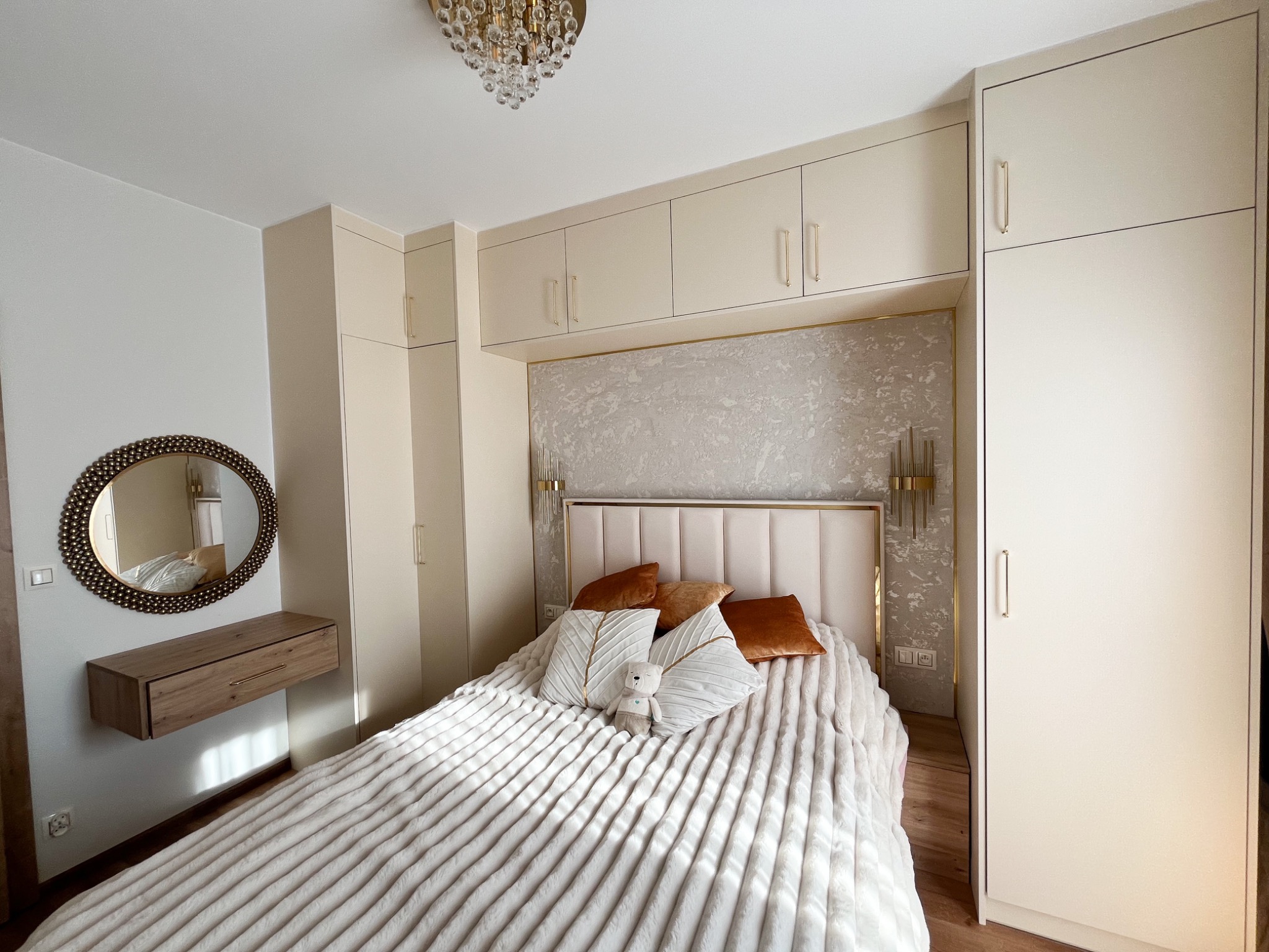 Beżowa sypialnia z szafami w zabudowie, łóżkiem z tapicerowanym zagłówkiem i wiszącą szafką nocną z lustrem w ozdobnej ramie. Całość oświetla kryształowy żyrandol.