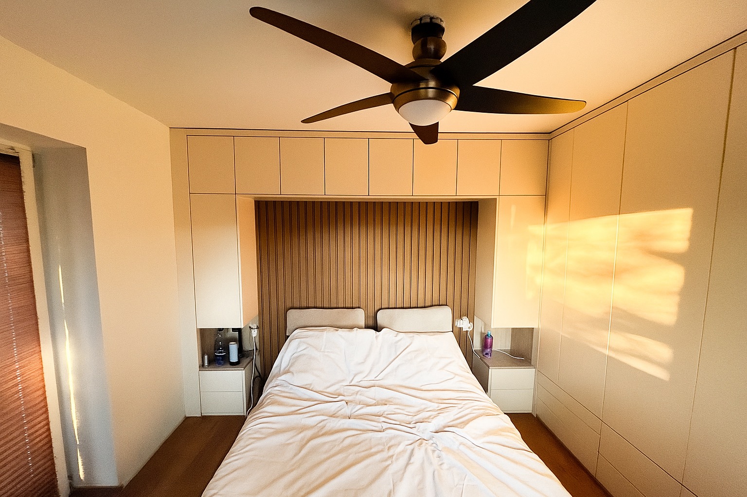 Sypialnia z łóżkiem i zabudową meblową z szafami po bokach i szafkami nad wezgłowiem. Drewniane panele dekoracyjne za łóżkiem. Oświetlenie sufitowe.