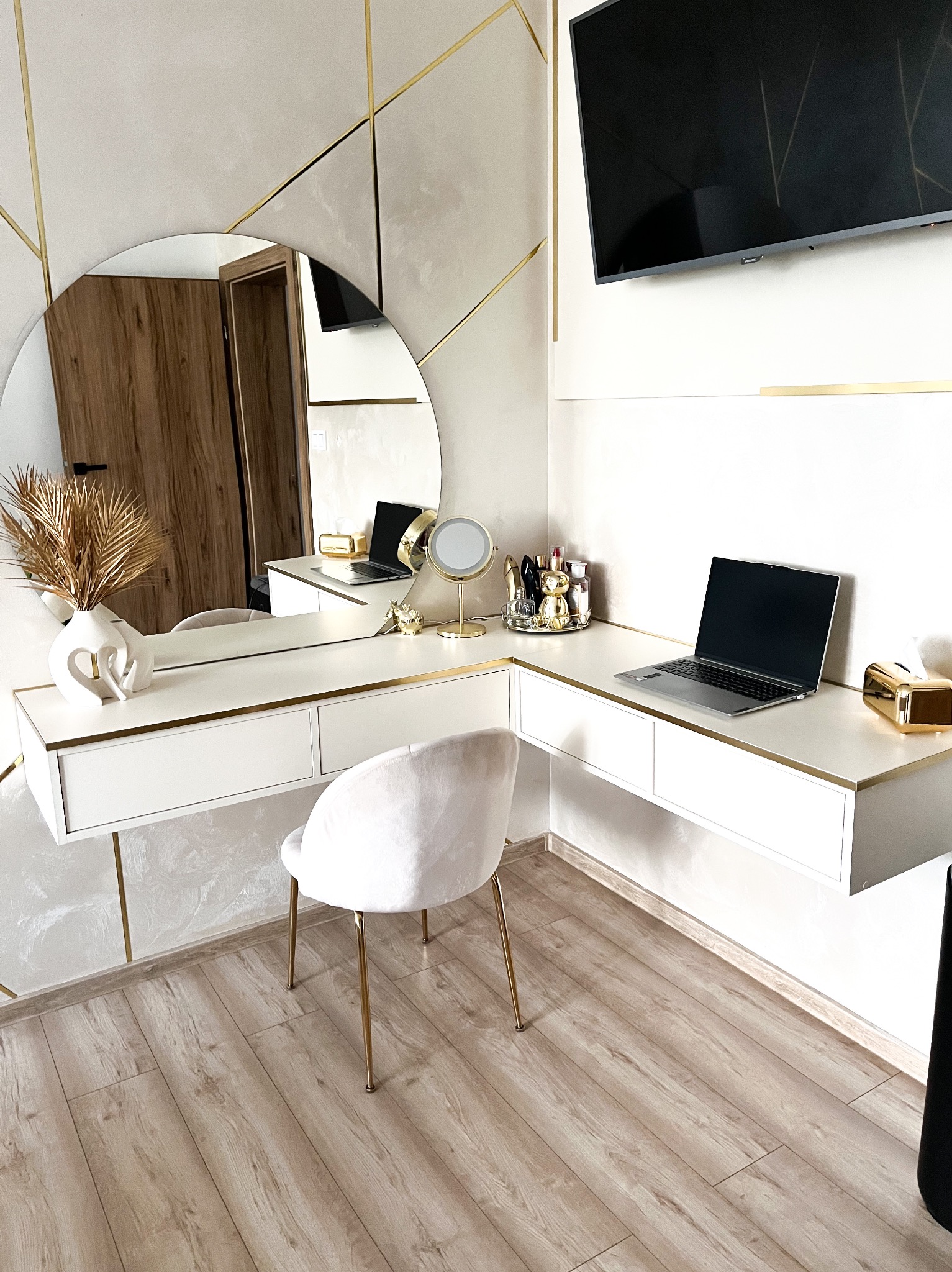 Biała toaletka z szufladami, okrągłym lustrem i złotymi akcentami. Na blacie laptop, wazon z suszem i kosmetyki. Krzesło z weluru na złotych nogach. Całość w jasnym, nowoczesnym wnętrzu.