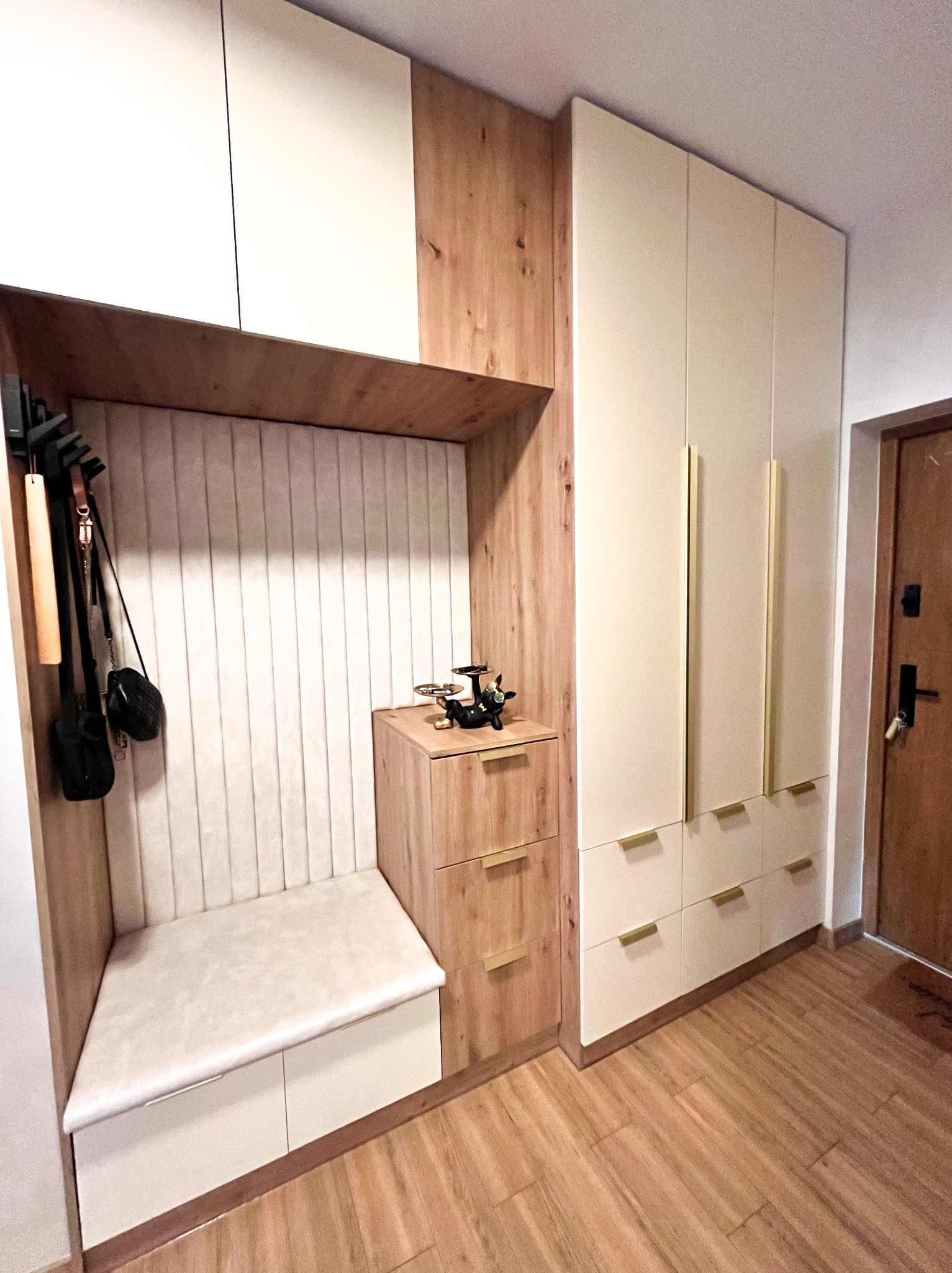 Funkcjonalna zabudowa przedpokoju z siedziskiem, wieszakami i szafą z jasnymi frontami i złotymi uchwytami. Drewnopodobne wykończenie dodaje ciepła.