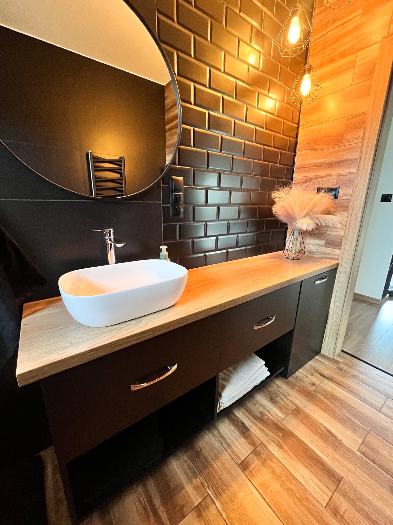 Nowoczesna łazienka z blatem drewnianym, umywalką nablatową i czarnymi płytkami w stylu metro. Uwagę przykuwają lampy wiszące i drewniane wykończenie obok drzwi.