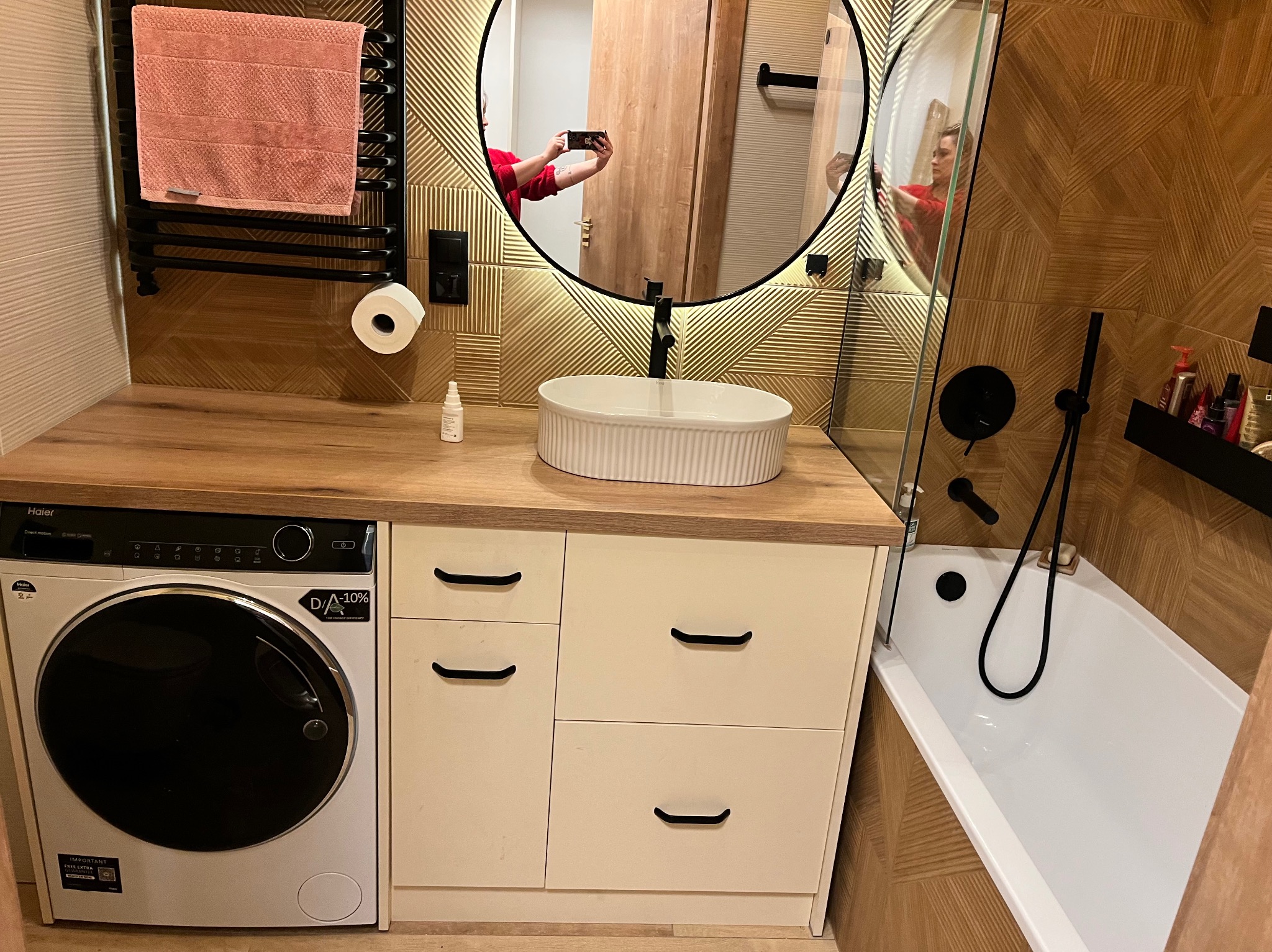 Nowoczesna łazienka z drewnianymi akcentami: umywalka nablatowa, pralka pod blatem, lustro w okrągłej ramie i mozaika na ścianie.