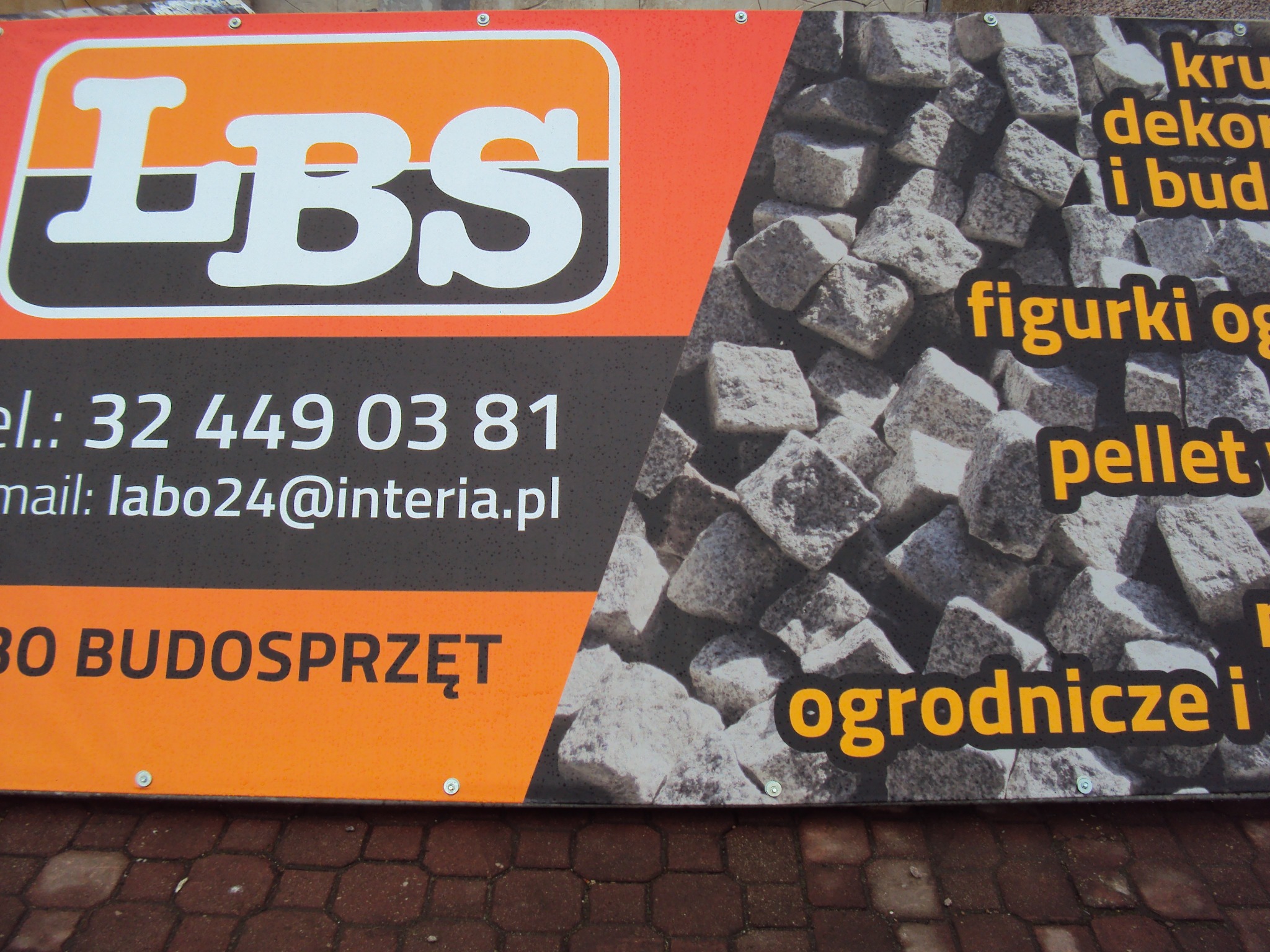 Baner reklamowy z logiem LBS Budosprzęt, prezentujący ofertę kruszyw dekoracyjnych i budowlanych, figurek ogrodowych i pelletu, z bliska widoczne nieregularne bryły szarego kamienia.