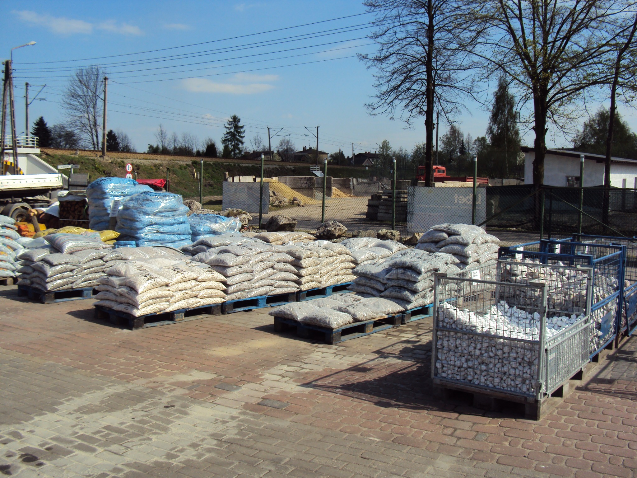 Składowisko materiałów budowlanych: palety z workami wypełnionymi jasnym kruszywem, niebieskie worki folii, metalowe kosze wypełnione białymi otoczakami, w tle budynki i drzewa.