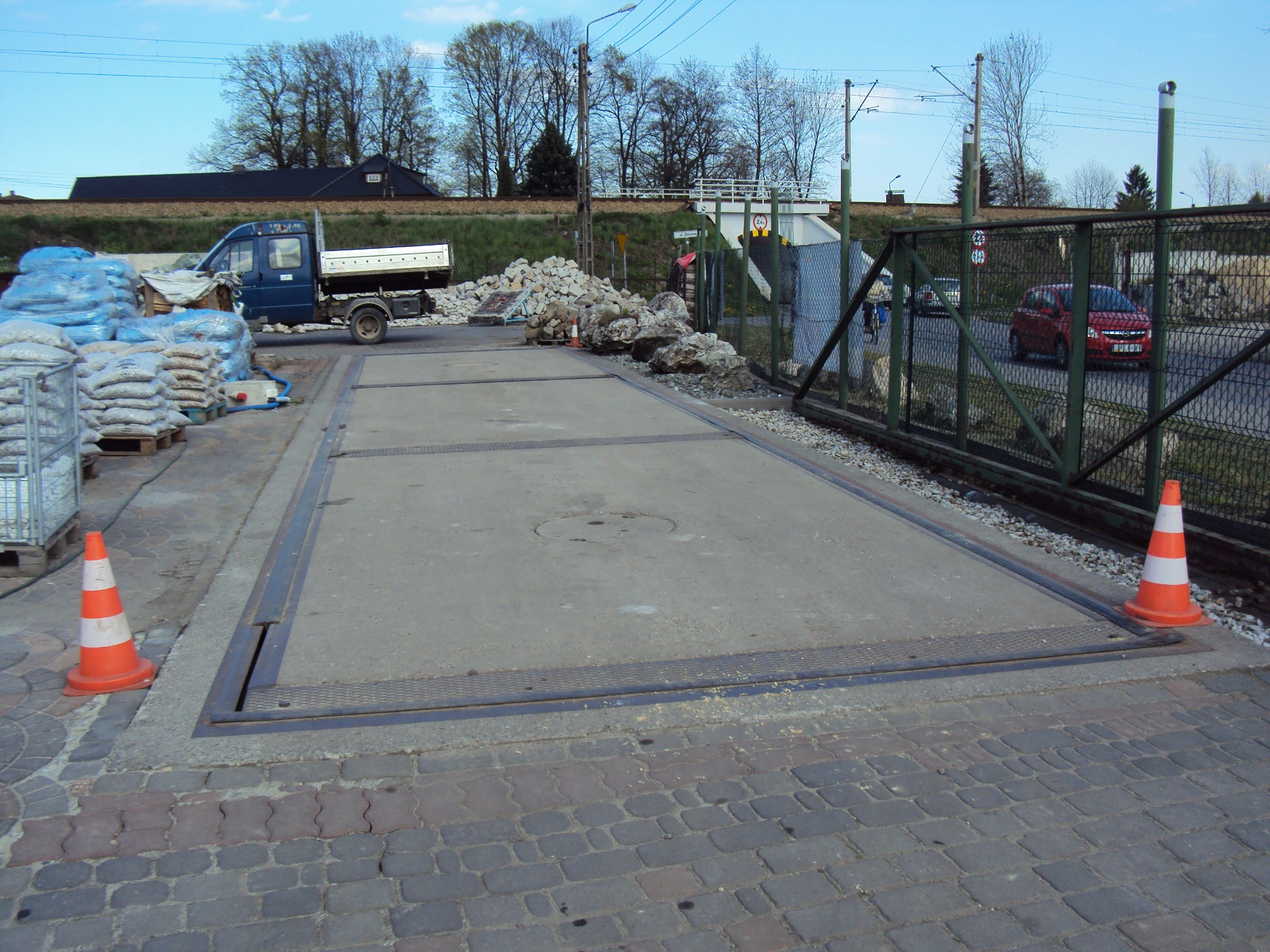 Waga samochodowa na placu budowlanym, obok zaparkowany samochód dostawczy z workami, w tle widoczne tory kolejowe i przejazd.