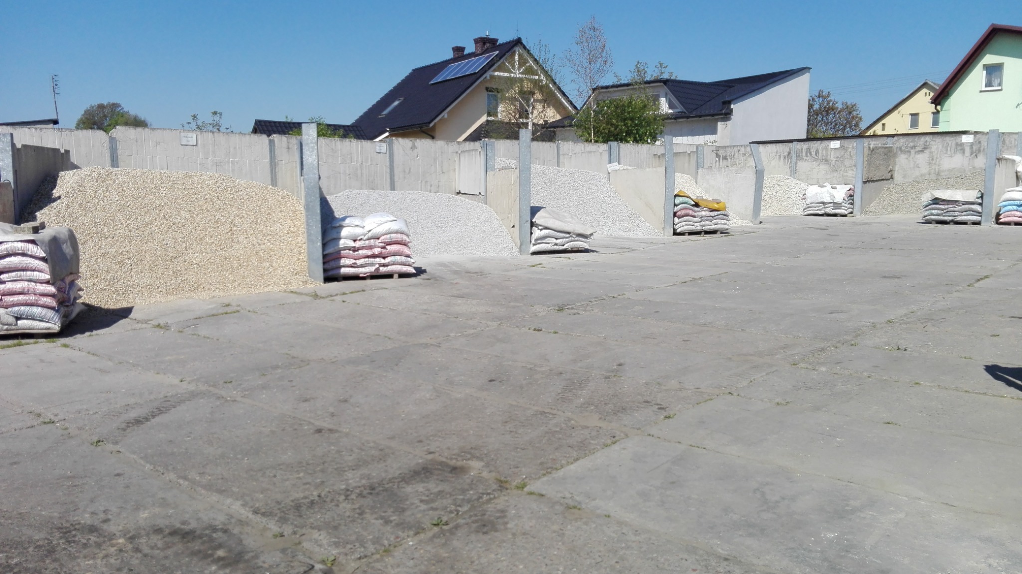 Składowisko kruszyw: pryzmy jasnego i szarego żwiru oraz piasku w workach, oddzielone betonowymi przegrodami, w tle zabudowa jednorodzinna.
