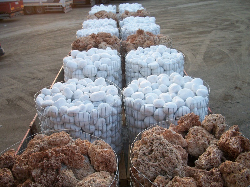 Rzędy metalowych koszy wypełnionych białymi, gładkimi otoczakami i porowatymi, brązowo-pomarańczowymi kamieniami na palecie transportowej.