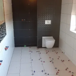 Łazienka w trakcie prac wykończeniowych: ułożone białe płytki podłogowe z krzyżykami dystansowymi, częściowo wykończona ściana z ciemnymi płytkami strukturalnymi, wisząca toaleta, widoczne przewody...