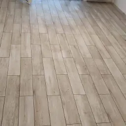 Ułożona podłoga z płytek imitujących drewno w jasnym odcieniu, widok na otwarte drzwi balkonowe zabezpieczone folią malarską i grzejnik na ścianie.