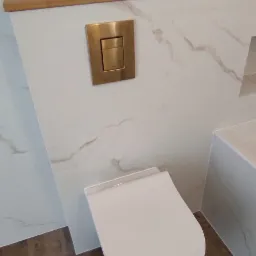 Nowoczesna toaleta podwieszana z białym siedziskiem i złotym przyciskiem spłukującym, marmurowe płytki ścienne i drewnopodobna podłoga.