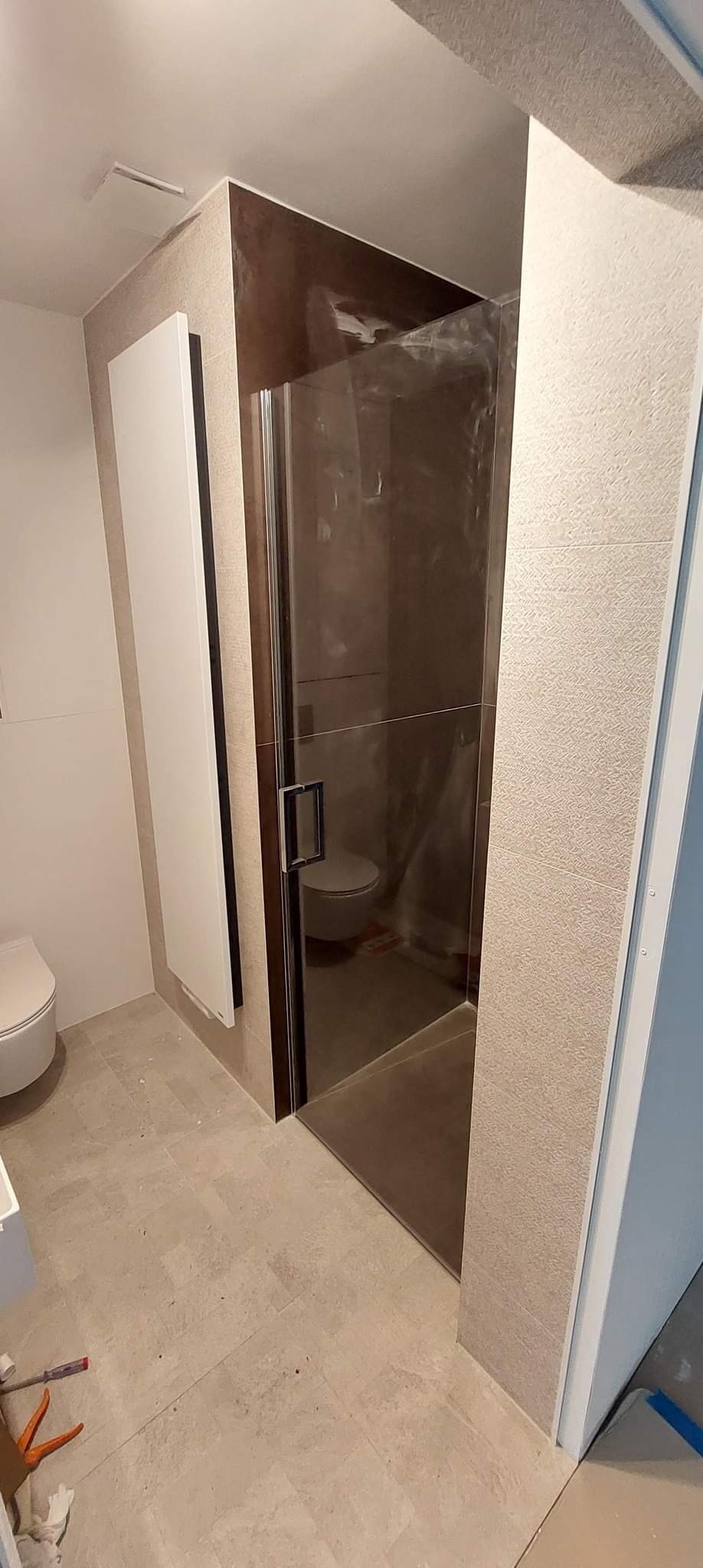 Nowoczesna kabina prysznicowa ze szklanymi drzwiami i brązowymi akcentami, widoczna toaleta w tle, jasne płytki podłogowe, grzejnik panelowy na ścianie, narzędzia na podłodze.