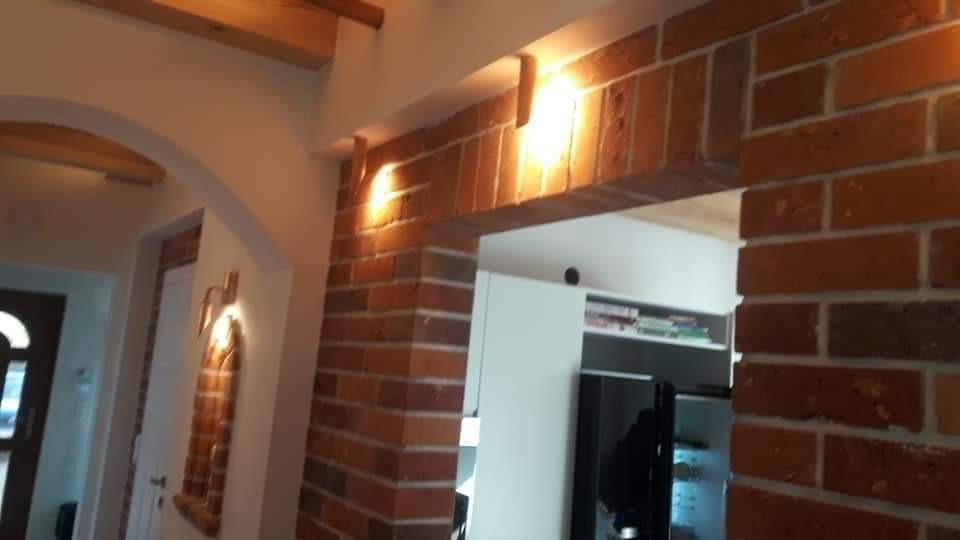 Ceglana ściana z oświetleniem punktowym, tworząca łukowate przejście do pomieszczenia z białymi meblami i czarną lodówką.