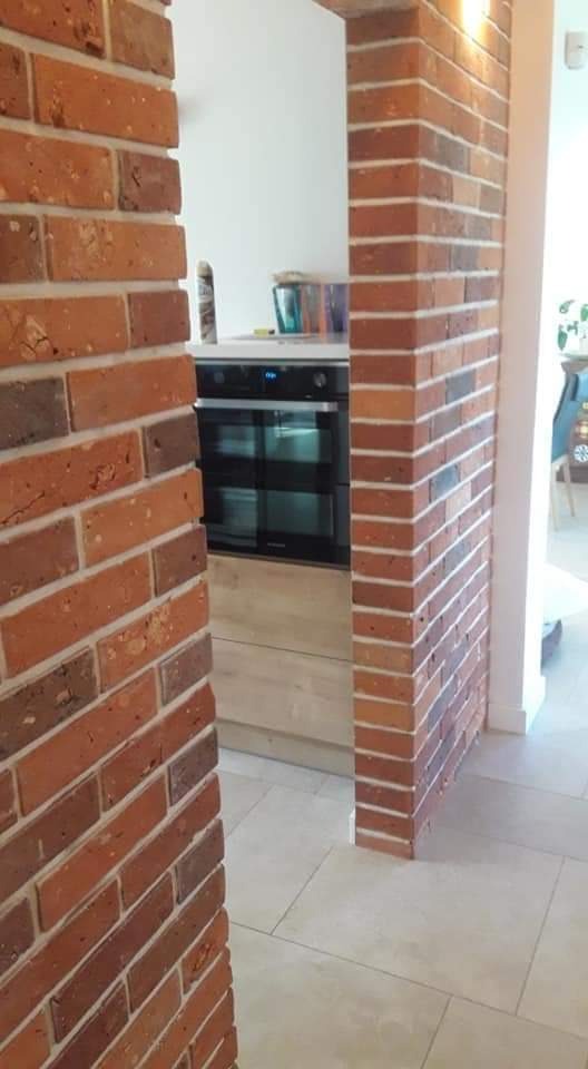 Ceglana kolumna oddzielająca kuchnię od salonu, widoczny fragment zabudowy kuchennej z piekarnikiem i jasne płytki podłogowe.