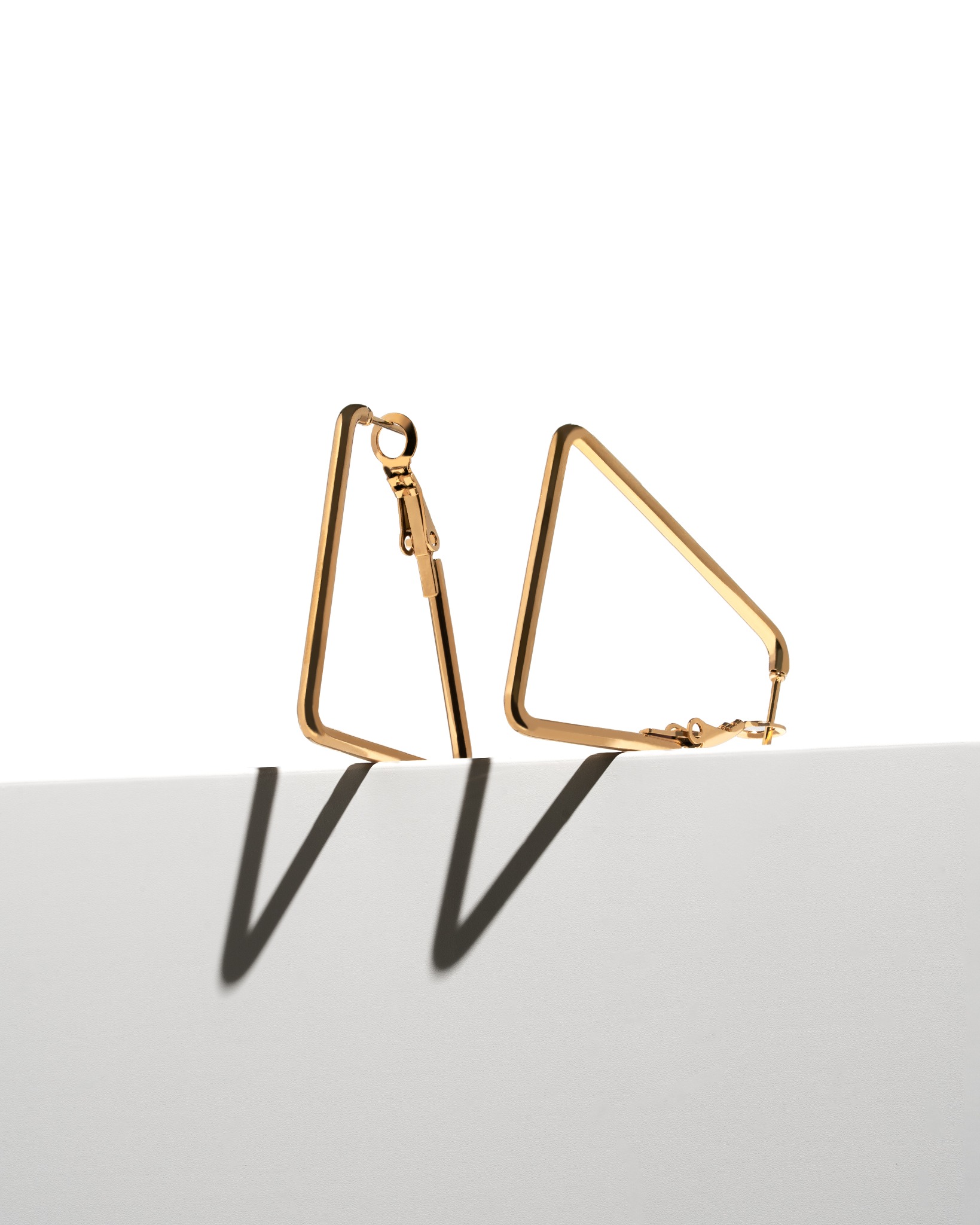 Dwa złote kolczyki w kształcie trójkątów na białym tle, rzucające wyraźny cień. Minimalistyczna fotografia produktowa biżuterii.