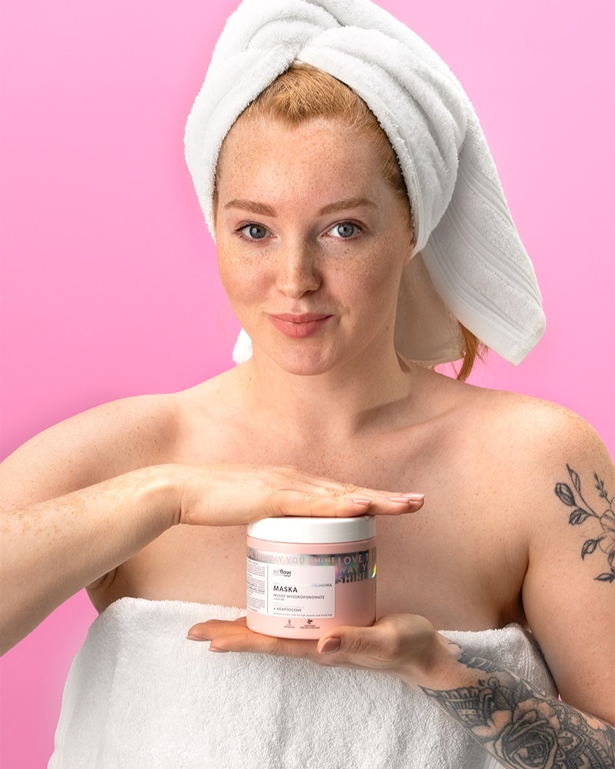 Uśmiechnięta kobieta z piegami, owinięta w biały ręcznik, prezentuje różową maskę do włosów na tle różowego studia. Widoczne tatuaże na ramionach.