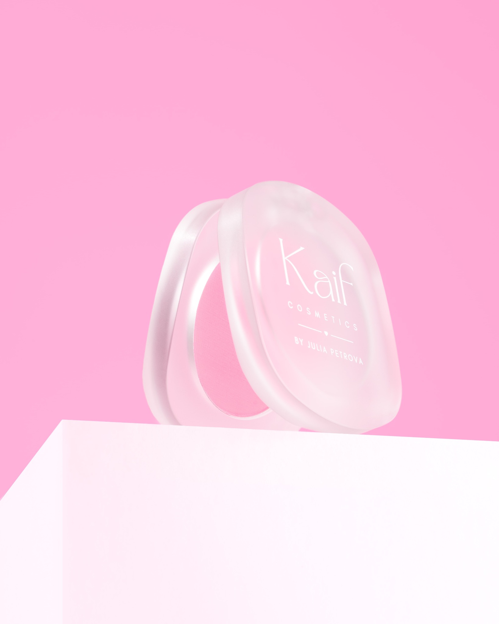 Puderniczka marki Kaif Cosmetics z otwartym wieczkiem, prezentowana na białym postumencie na tle różowego studia. Makro, delikatne światło.