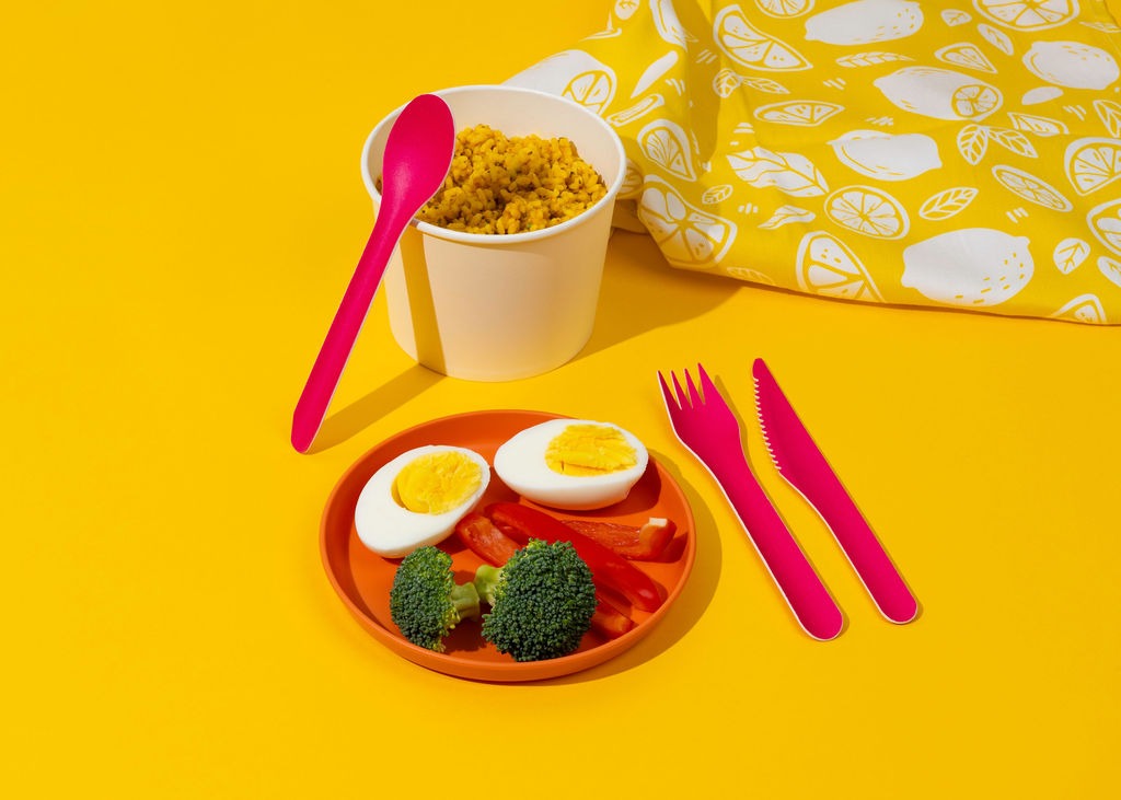 Żółte tło z posiłkiem w misce, na talerzu jajko, brokuły, papryka i różowe sztućce. W tle torba z motywem cytryn. Zdjęcie produktowe.