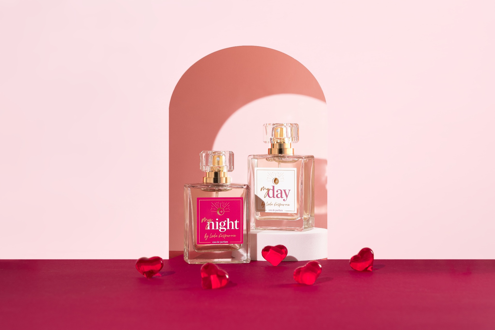 Dwa flakony perfum 'My Night' i 'My Day' na białym postumencie, otoczone czerwonymi sercami na burgundowym tle, na tle różowej ściany z łukiem.