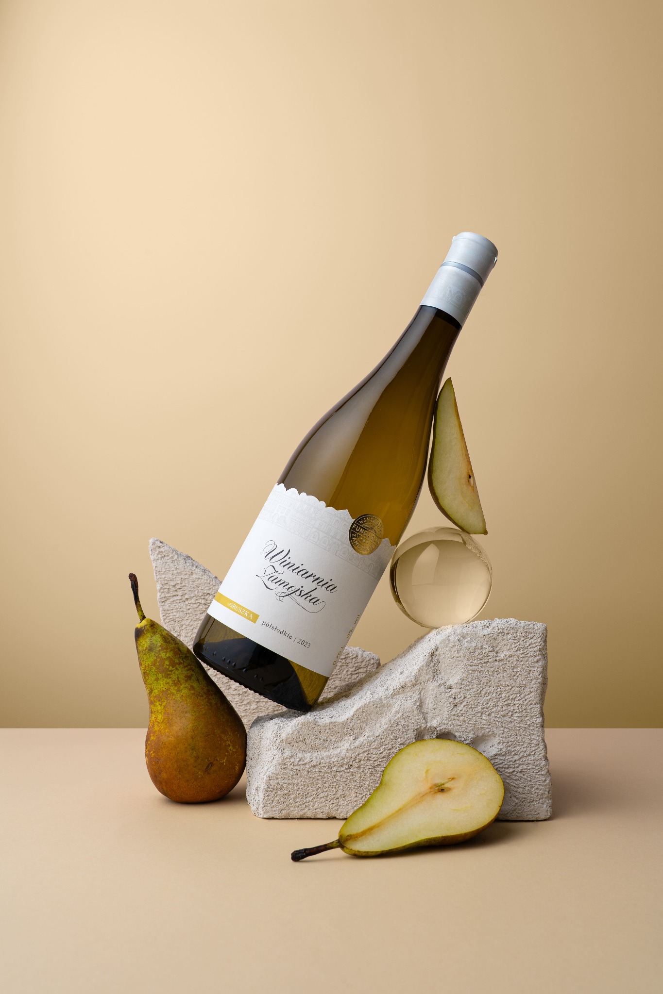 Artystyczne ujęcie butelki białego wina 'Winiarnia Zamojska' z gruszkami i elementami dekoracyjnymi na beżowym tle. Nietypowa kompozycja, jasne tło, dobre oświetlenie.