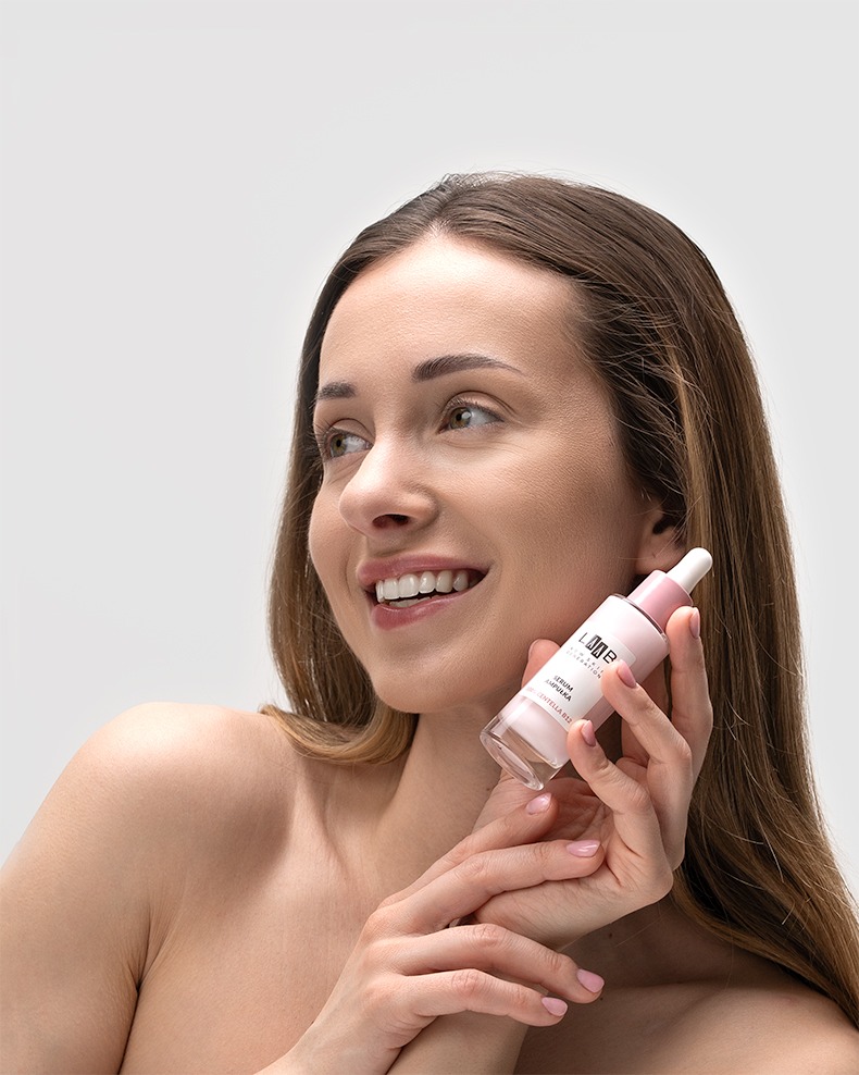 Uśmiechnięta kobieta z serum do twarzy w rękach, jasne tło, subtelne oświetlenie. Produkt marki IMAGE, delikatny różowy odcień. Promienna cera.