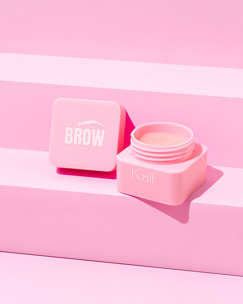 Różowe kosmetyki do brwi marki Kaif Cosmetics na tle w pastelowych odcieniach różu. Pudełko mydła do brwi i otwarty słoiczek balsamu. Minimalistyczna fotografia produktowa.