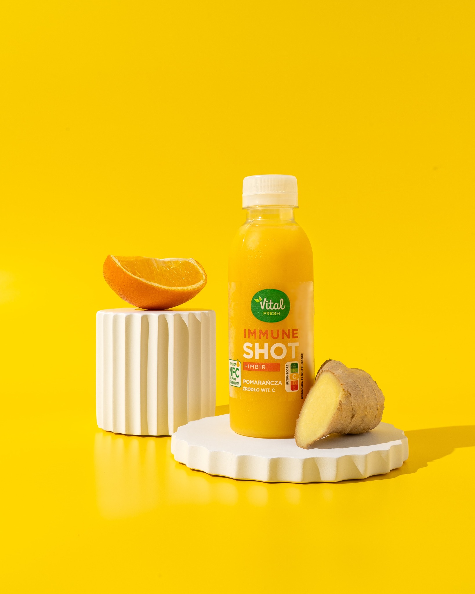 Żółty napój w butelce Vital Fresh Immune Shot z imbirem i pomarańczą, na białych postumentach, na żółtym tle. Fotografia produktowa.