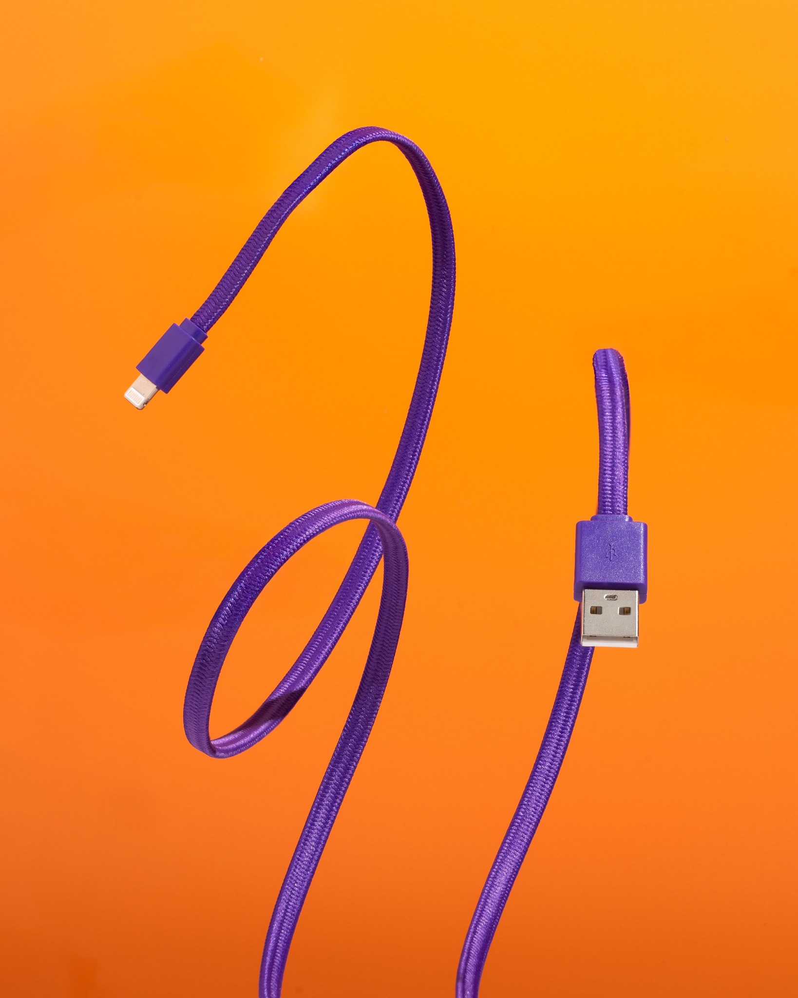 Fioletowy kabel USB z oplotem na pomarańczowym tle. Widoczne złącza USB-A i Lightning. Kabel ułożony w kształt fali. Zdjęcie produktowe.
